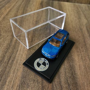 May include: A blue die-cast model car on a black base with the BMW logo. The miniature car is displayed inside a clear acrylic box. The car has a detailed interior and exterior design.