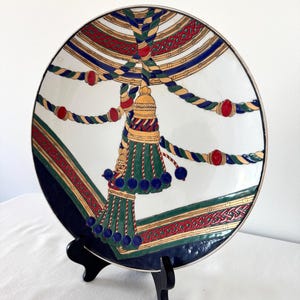 Puede incluir: Un plato blanco decorativo con un dise&ntilde;o detallado con detalles en verde, dorado, rojo y azul. El plato tiene un dise&ntilde;o de borla y cuerda, con un borde azul marino. El plato se exhibe sobre un soporte negro.
