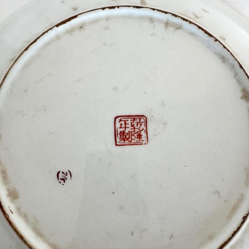 Puede incluir: La parte inferior de un plato de cer&aacute;mica blanco con un borde marr&oacute;n. Un sello cuadrado rojo con caracteres chinos est&aacute; centrado en el plato. Una marca roja m&aacute;s peque&ntilde;a est&aacute; a la izquierda del sello.