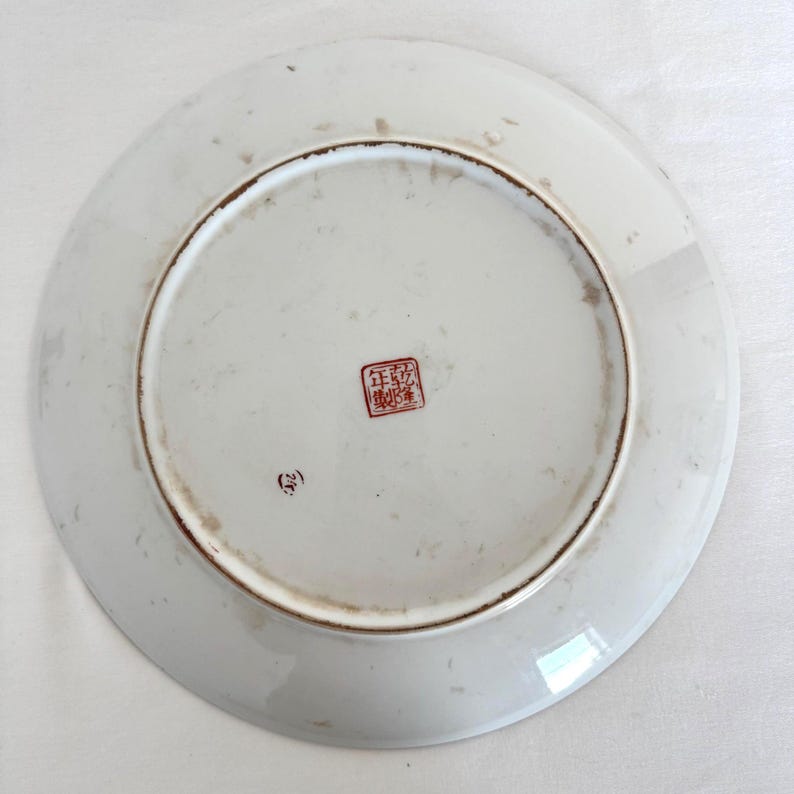 Puede incluir: Plato de cer&aacute;mica blanco con borde marr&oacute;n. La parte inferior del plato presenta un sello rojo con caracteres asi&aacute;ticos y un peque&ntilde;o n&uacute;mero entre par&eacute;ntesis. El plato tiene un dise&ntilde;o circular.