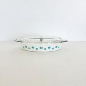 May include: White oval casserole dish with a clear glass lid. The dish features a turquoise snowflake pattern around the rim. The handles are clear glass. A vintage kitchenware item.