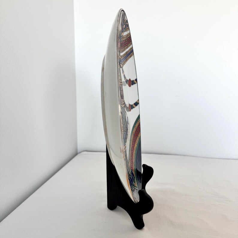 Puede incluir: Un plato de cer&aacute;mica blanco con un borde decorativo que presenta un dise&ntilde;o colorido de patrones rojos, azules y dorados. El plato se exhibe sobre un soporte negro, mostrando su perfil lateral sobre un fondo blanco.