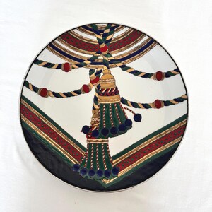 Puede incluir: Un plato decorativo con un centro blanco y un dise&ntilde;o detallado. El dise&ntilde;o presenta una borla dorada con detalles en verde y azul, cuerda roja y dorada y un borde rojo y dorado. El plato tiene un borde negro.