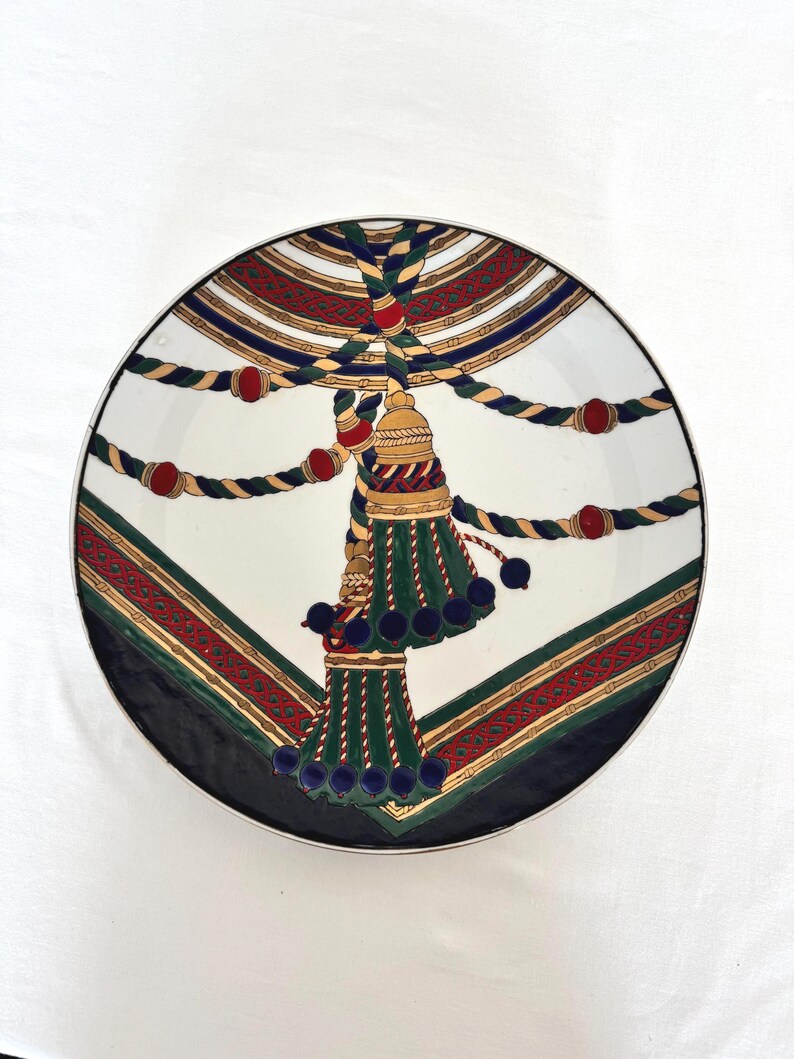 Puede incluir: Un plato decorativo con un centro blanco y un dise&ntilde;o detallado. El dise&ntilde;o presenta una borla dorada con detalles en verde y azul, cuerda roja y dorada y un borde rojo y dorado. El plato tiene un borde negro.