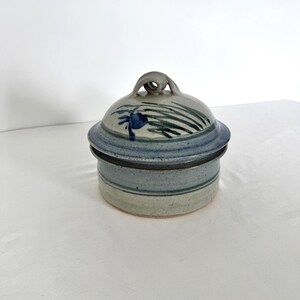 May include: A ceramic lidded container with a round base and a domed lid. The lid features a loop handle and is decorated with blue and green painted floral designs. The container is primarily blue and gray, with a light-colored base.