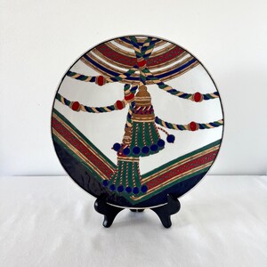 Puede incluir: Un plato decorativo de cer&aacute;mica con un dise&ntilde;o de borla. El plato presenta un fondo blanco con un motivo central de borla en verde, dorado y azul. El borde tiene un patr&oacute;n rojo y verde con un borde azul marino. El plato se exhibe en un soporte negro.