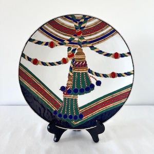 Puede incluir: Un plato decorativo blanco con borde plateado, con un dise&ntilde;o detallado de borla y motivo de cuerda. El dise&ntilde;o incorpora colores rojo, verde, azul y dorado. El plato se exhibe sobre un soporte negro.