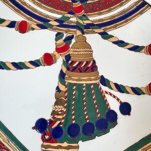 Puede incluir: Primer plano de un plato decorativo con un dise&ntilde;o detallado. El dise&ntilde;o presenta una borla central en verde, azul y dorado, con detalles de cuerda roja y azul. El fondo es blanco y el estilo general es ornamentado.