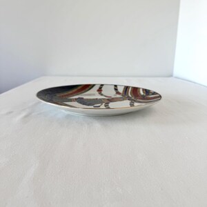 Puede incluir: Un plato de cer&aacute;mica decorativo con borde dorado. El plato presenta un dise&ntilde;o colorido con patrones rojos, azules y marrones. El plato est&aacute; sobre una superficie blanca.