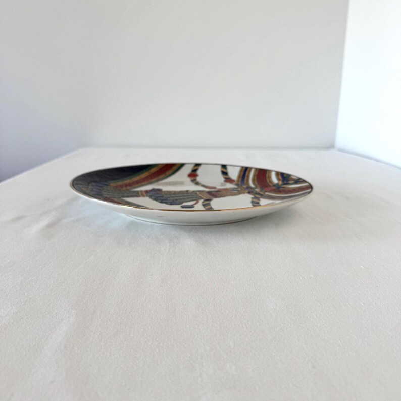 Puede incluir: Un plato de cer&aacute;mica decorativo con borde dorado. El plato presenta un dise&ntilde;o colorido con patrones rojos, azules y marrones. El plato est&aacute; sobre una superficie blanca.