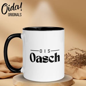 Könnte beinhalten: Weißer Keramikbecher mit schwarzem Innenraum und Henkel. Der Becher trägt den Text "-OIS- Oasch" in Schwarz, mit dem Markennamen "Oida! ORIGINALS" in schwarzer Schrift oben. Der Becher steht auf einem Holzuntersetzer.