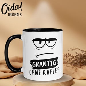 Op de afbeelding: Witte keramische mok met een zwart handvat en rand, met een chagrijnig gezichtsontwerp. De mok heeft de tekst "GRANTIG OHNE KAFFEE" in het zwart. De mok staat op een houten standaard.