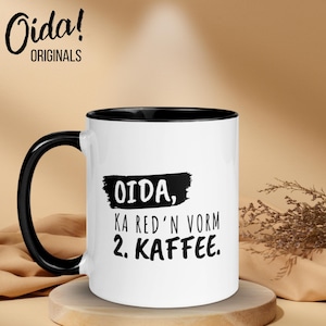 Pode incluir: Caneca de café de cerâmica branca com alça e borda pretas. A caneca apresenta o texto "OIDA, KA RED'N VORM 2. KAFFEE." em preto, com a marca "Oida! ORIGINALS" no canto superior esquerdo.