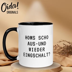 Może przedstawiać: Biały ceramiczny kubek z czarnym uchwytem i wnętrzem. Kubek zawiera tekst "HOMS SCHO AUS-UND WIEDER EINGSCHALT?" w pikselowanej czcionce. Kubek ma logo "Oida! ORIGINALS".