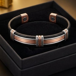 May include: A cuff bracelet featuring a black leather band accented with central copper and silver-toned metal strips. The bracelet has metallic end caps with visible circular magnets and is presented inside a square black box.