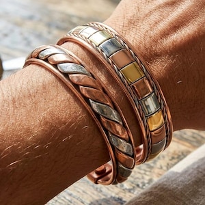 Può includere: Due bracciali rigidi in metallo indossati insieme. Un bracciale presenta un design a corda intrecciata in toni alternati di rame e argento. Il secondo bracciale mostra una serie di segmenti quadrati con finiture metalliche alternate in rame, argento e oro.