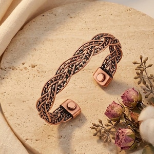Könnte beinhalten: Ein kupferfarbenes Metall-Armband mit einem komplexen geflochtenen Drahtdesign. Das Armband weist ein dunkles, oxidiertes Finish innerhalb des gewebten Musters auf und enthält zwei kleine, kreisförmige magnetische Elemente an den offenen Enden.
