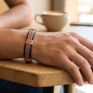 May include: A cuff bracelet featuring a central copper-toned metal band with a repeating wave-like pattern, flanked by two smooth black leather-like strips. The design is secured with copper-toned metal accents at the centre and the edges.
