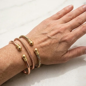 Puede incluir: Un conjunto de tres pulseras abiertas tipo brazalete en la muñeca. Una pulsera es de metal liso en tono cobre, mientras que las otras dos presentan una textura trenzada. Cada extremo de los brazaletes termina en una bola de metal dorado pulido.