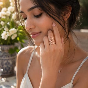 Könnte beinhalten: Ein roségoldener Ring mit einem doppelten Wirbeldesign. Der Ring befindet sich an einem Finger, und die Person trägt ein weißes Kleid. Eine zarte Halskette mit einem kleinen Anhänger ist ebenfalls sichtbar.