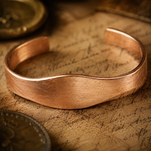May include: A copper cuff bracelet with a brushed, matte finish and a slightly curved, wave-like top edge. The bracelet is an open-ended band with a smooth, metallic surface that reflects light, showcasing a warm, reddish-brown colour throughout.