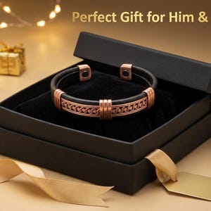 Puede incluir: Una pulsera de cuero negro con detalles metálicos de color cobre con un patrón trenzado y dos imanes circulares en los extremos abiertos, presentada dentro de una caja negra con el texto Perfect Gift for Him & Her impreso arriba.