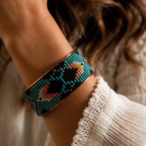 May include: A wide cuff bracelet featuring a geometric pattern of small glass beads in shades of turquoise, black, and peach. The bracelet has a metallic copper-toned frame that borders the intricate beadwork, creating a structured design.