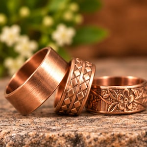 May include: Three copper-coloured rings arranged in a row. The first ring on the left has a smooth, brushed finish. The middle ring features a repeating geometric diamond pattern. The third ring on the right displays an embossed floral design.