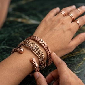 Könnte beinhalten: Ein Set aus drei kupferfarbenen Armbändern am Handgelenk, bestehend aus einer geflochtenen Manschette, einem gewebten Band und einer strukturierten Gliederkette. Vier dünne kupferfarbene Ringe befinden sich an den Fingern derselben Hand.