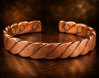 Puede incluir: Una pulsera tipo brazalete de color cobre con un diseño de cuerda trenzada y un acabado texturizado y desgastado. El interior de la banda abierta muestra varios pequeños insertos magnéticos circulares de tono metálico incrustados.