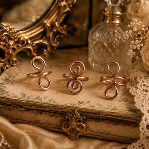 Puede incluir: Tres anillos de metal en tono oro rosa, cada uno con un diseño de alambre continuo formado por dos bucles o espirales interconectados. Los anillos están dispuestos en una fila horizontal, mostrando un acabado metálico pulido y una estructura de banda simple.