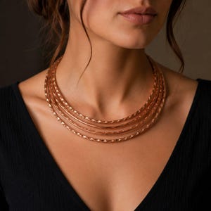 May include: A multi-strand necklace featuring four distinct circular bands in a warm rose gold tone. Each band displays a textured, twisted metal finish that catches the light, creating a layered appearance around the neck area.