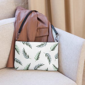 Könnte beinhalten: Weiße Clutch mit schwarzem Riemen und einem sich wiederholenden Muster aus grünen und grauen Farnblättern. Die rechteckige Tasche hat einen Reißverschluss. Im Hintergrund ist ein brauner Blazer zu sehen.