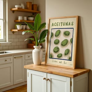 Op de afbeelding: Een ingelijste print met het woord "ACEITUNAS" en illustraties van groene olijven. De print zit in een houten lijst en staat op een witte kast met een houten blad. Een witte vaas met een groene plant staat ernaast.