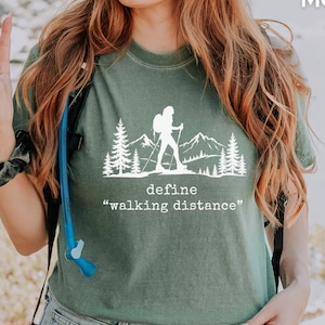 Può includere: Una maglietta verde muschio con una grafica bianca di un escursionista con montagne e alberi, e la scritta "define walking distance". La maglietta è realizzata in un materiale morbido.