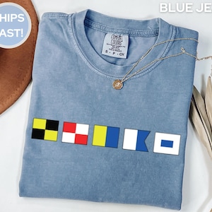 May include: A blue jean colored t-shirt with nautical flag graphics. The shirt has a crew neck and short sleeves. A gold necklace with a pendant is draped across the shirt. The text "BLUE JEAN" is at the top.