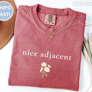 May include: A crimson t-shirt with the words "nice adjacent" printed in white. A small floral graphic is below the text. A silver necklace with a pendant rests on the shirt.