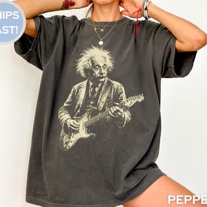 May include: A charcoal gray short-sleeve t-shirt featuring a cream-colored graphic print of Albert Einstein playing an electric guitar. The shirt has a crew neckline and a relaxed fit, with the word PEPPER printed in the bottom right corner.