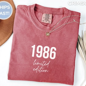 May include: Crimson-colored t-shirt with the year "1986" printed in white, along with the words "limited edition" in a script font. A delicate silver necklace with a pendant rests on the shirt. The shirt is folded neatly.