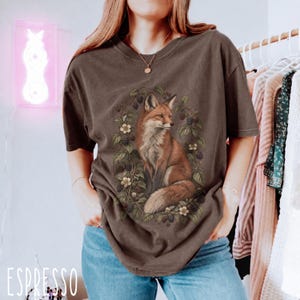 Peut inclure: Un t-shirt à manches courtes marron foncé avec un motif central représentant un renard roux assis au milieu d'une couronne de feuilles vertes, de fleurs blanches et de mûres sombres. Le t-shirt a un col rond et une coupe décontractée.