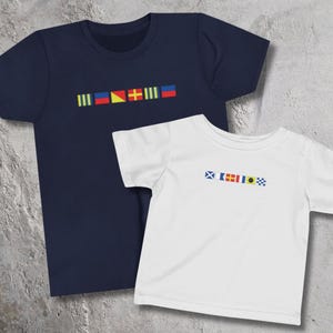 May include: A navy blue t-shirt and a white t-shirt, each featuring a horizontal row of colorful nautical signal flags printed across the chest. The navy shirt displays six distinct flags, while the white shirt displays seven smaller flags.