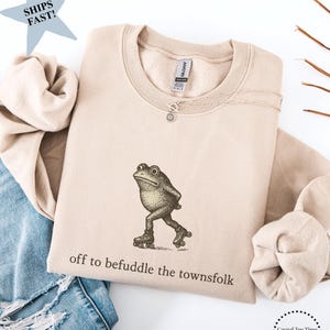 Könnte beinhalten: Beigefarbenes Sweatshirt mit einer Vintage-Illustration einer Frosch auf Rollschuhen. Der Text "off to befuddle the townsfolk" ist darunter gedruckt. Das Sweatshirt hat einen Rundhalsausschnitt und ist aus weichem Material.