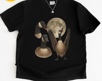 Op de afbeelding: Zwart T-shirt met een afbeelding van ganzen en een volle maan. De ganzen worden in verschillende poses afgebeeld. Op de onderkant van het shirt staat de tekst "black" en "Casual Tee Time".