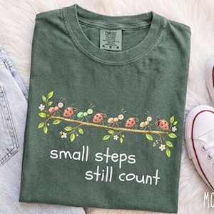 May include: A dark olive green t-shirt featuring a graphic of ants and ladybugs walking along a leafy branch. Below the illustration, the white text reads, "small steps still count." The shirt has a crew neckline and a Comfort Colors brand label.