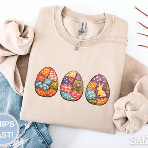 May include: A sand-coloured sweatshirt with three embroidered Easter eggs. The eggs feature patchwork designs with colourful patterns, including flowers and a bunny. A silver necklace with a small pendant is visible. The word "SAND" is printed on the bottom right.