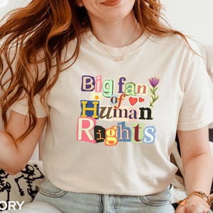 Może przedstawiać: Kremowa koszulka z napisem "Big fan of Human Rights" w kolorowym, wyciętym stylu. Projekt zawiera serca i kwiatowe akcenty. Koszulka jest wykonana z miękkiego materiału.