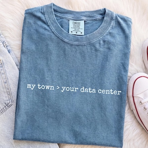 Peut inclure: Un t-shirt bleu à manches courtes avec un col rond et une étiquette de marque Comfort Colors à l'intérieur du col. Le t-shirt présente le texte imprimé en blanc "my town > your data center" centré sur la poitrine dans une police simple.