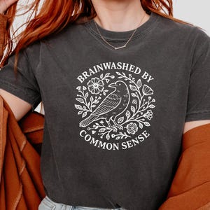 Op de afbeelding: Donkergrijs t-shirt met een witte afbeelding. De afbeelding toont een raaf in een bloemenkrans met de woorden "BRAINWASHED BY COMMON SENSE". Het shirt heeft korte mouwen en een ronde hals.