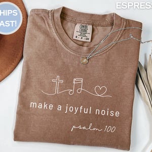 Peut inclure: Un t-shirt à manches courtes de couleur espresso avec un graphisme en ligne blanche représentant une croix, une note de musique et un cœur. Sous le graphisme, le texte "make a joyful noise" et "psalm 100" est écrit en blanc.