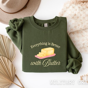 May include: Military green sweatshirt with the phrase "Everything is better with Butter" and a graphic of butter on a pink cutting board. The text and graphic are in a light yellow color.